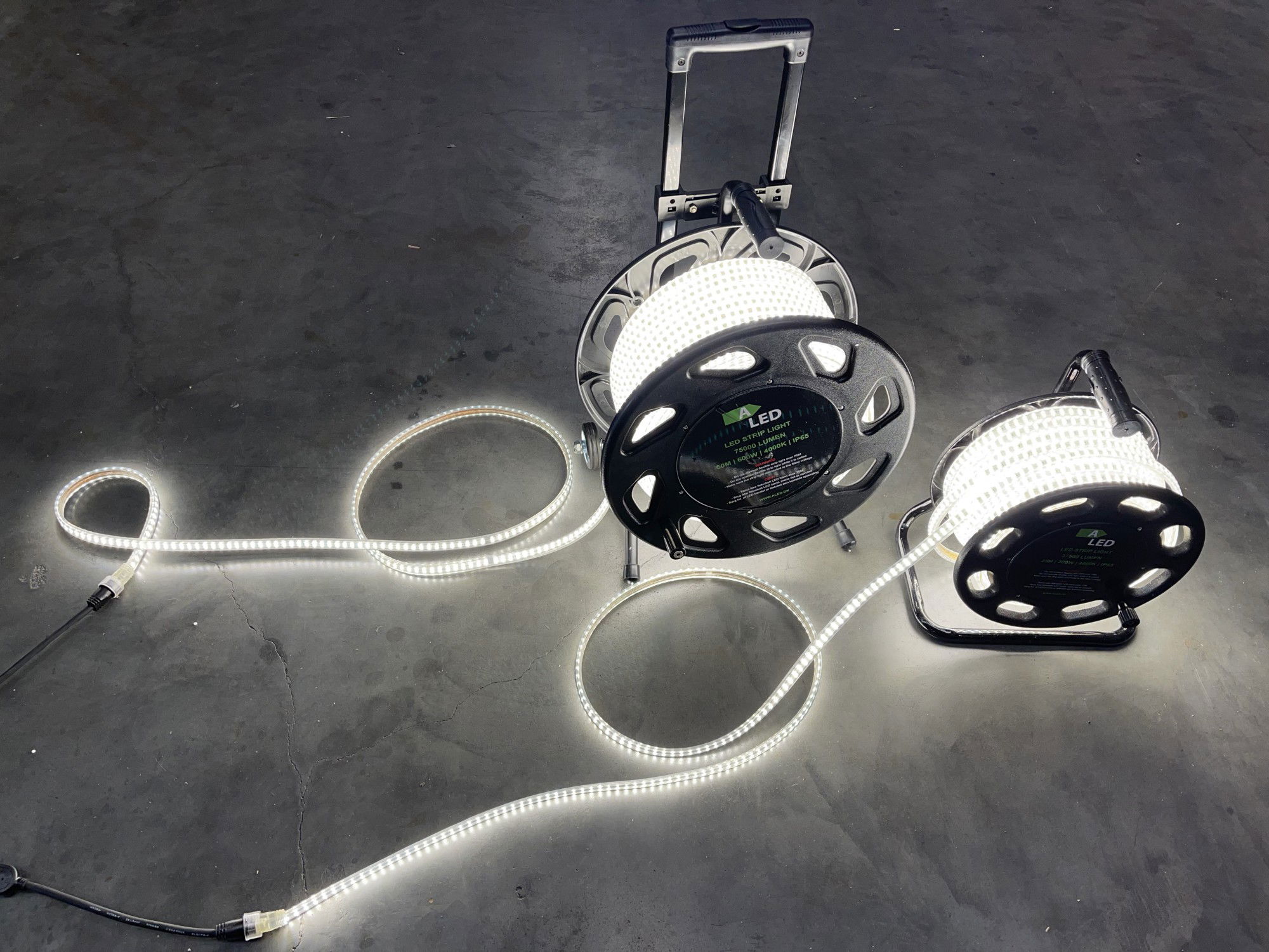 LED strip trumma 50m, 230V med 1500lm per meter, 4000K neutralvitt och IP65 skyddsklass. Två svarta trummor står på golvet med utrullat och tänt LED band som ger stark arbetsbelysning.