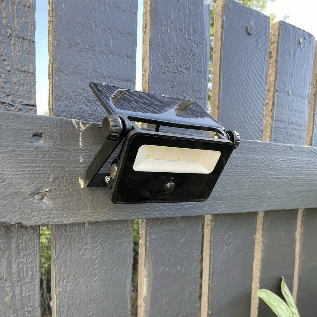 Sort solcelle projektør med 8 SMD LED dioder og indbygget solpanel, monteret på en grå træhegn til udendørs belysning.