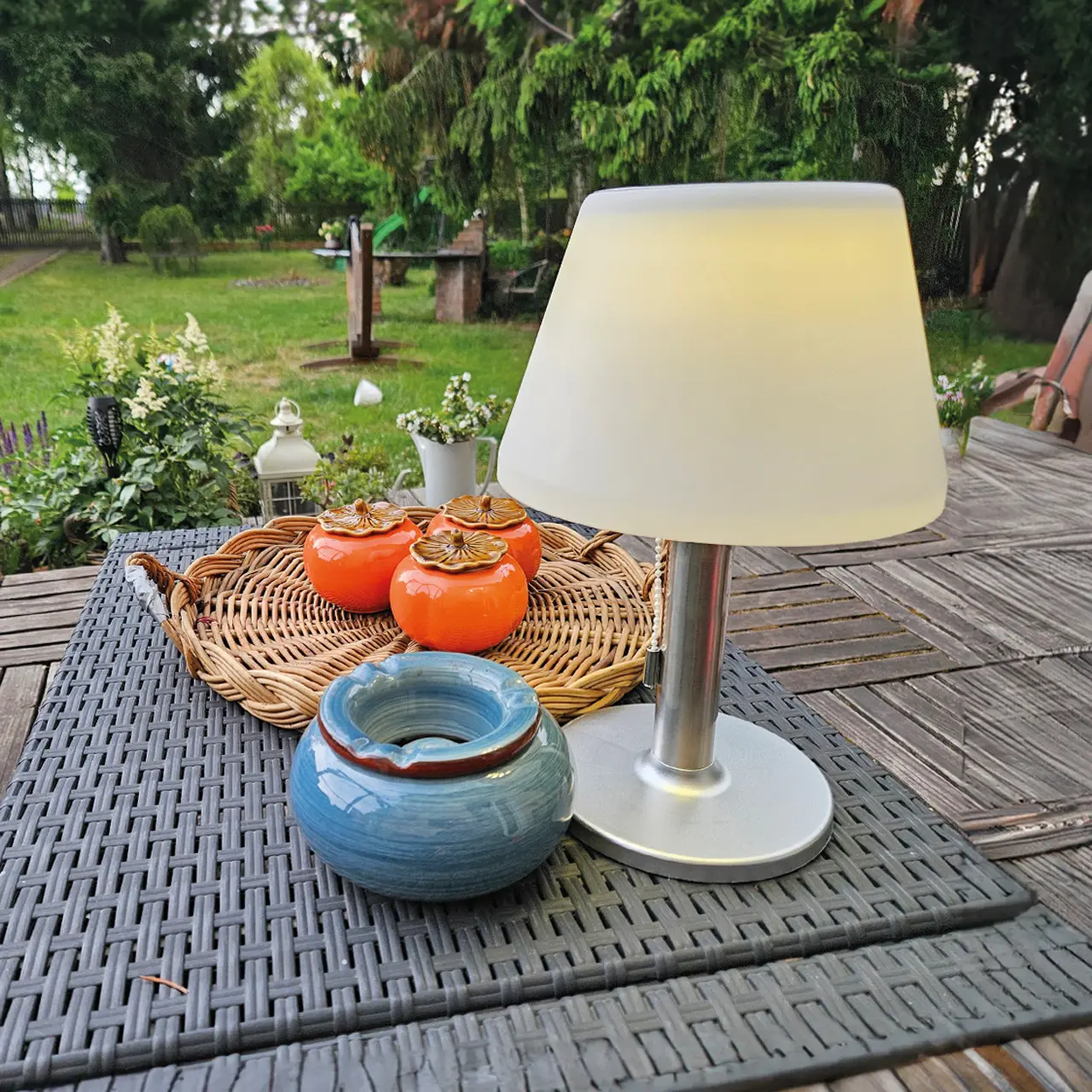 Skrivbordslampa i rostfritt stål med vit lampskärm, stående på ett terrassbord i dagsljus bredvid en blå keramikskål och en flätad bricka med dekorativa burkar.