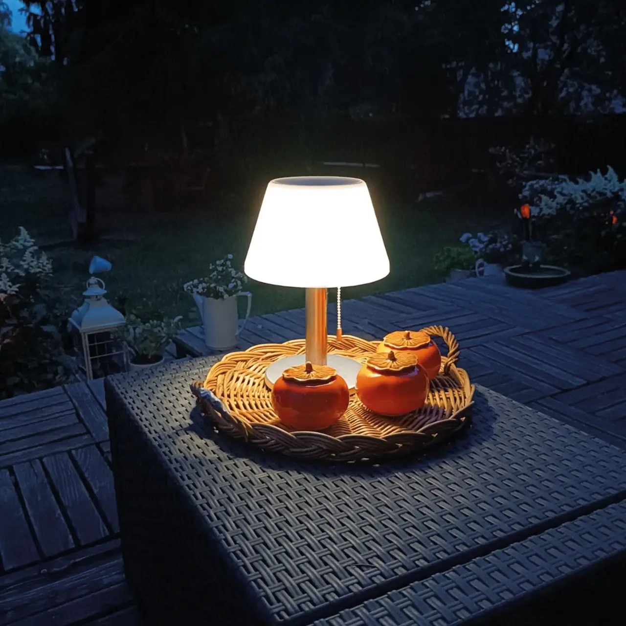 Kompakt skrivbordslampa i krom och vit plast med inbyggd LED, placerad på ett utomhusbord på kvällen och lyser över en flätad bricka med orangefärgade burkar.